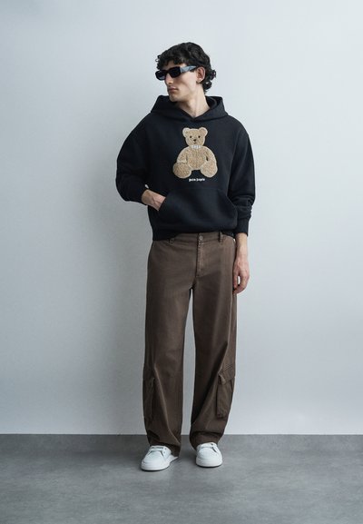 Jonge man die een zwarte hoodie met teddybeerafbeelding draagt, bruine cargobroek, witte sneakers en zwarte zonnebril, staand tegen een grijze achtergrond.