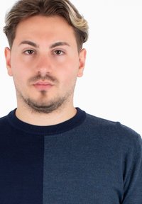 Młody mężczyzna z krótkimi falującymi włosami, ubrany w sweter w granatowo-ciemnoniebieski kolor w podziale, z neutralnym wyrazem twarzy, na białym tle.