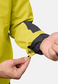 Giacca impermeabile con un corpo giallo brillante, dettaglio nero sulla manica, polsini elasticizzati e chiusura a scatto ai polsi.