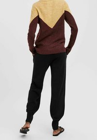 Maglione lavorato a maglia con un design a blocchi di colore giallo e bordeaux, abbinato a pantaloni della tuta neri a gamba stretta e ciabatte nere. Texture morbida.