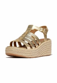 Sandales à talons compensés dorées avec une plateforme en jute tressé. Présente des brides entrelacées et une fermeture à boucle au niveau de la cheville. Texture lisse.