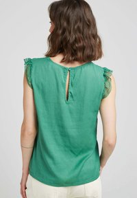Haut sans manches vert, présentant une finition en dentelle sur les manches, une fermeture arrière en forme de trou de serrure avec un bouton, et un tissu texturé.
