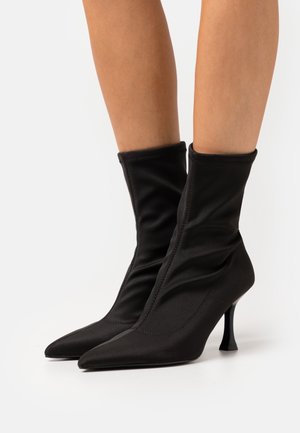 Botines negros de tobillo hechos de tela suave, con un diseño arrugado, punta afilada y un tacón delgado y estilizado.