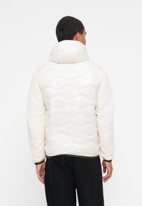 Veste blanche rembourrée avec capuche, dotée d'un design matelassé ondulé, de poignets élastiques et d'un ourlet droit. Texture lisse et silhouette moderne.