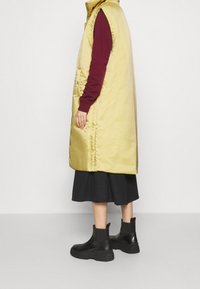 Gul vadderad ärmlös jacka med hög krage, bärs över en vinröd långärmad topp, kombinerad med en svart kjol och kraftiga svarta ankelstövlar.