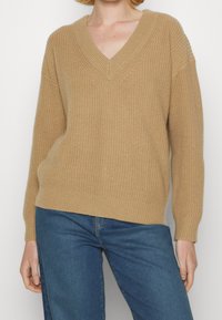 Pull beige col en V avec texture côtelée et manches larges, associé à un jean bleu. Le tissu en maille présente des points visibles et une coupe décontractée.