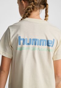 Lys beige t-shirt med korte ærmer, der har et stort blåt "hummel" logo og grønt "PERFORMANCES" tekst på bagsiden. Blødt stof.