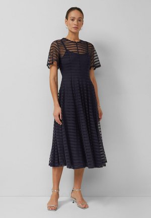 Frau in einem marineblau gestreiften, durchsichtigen Midikleid mit kurzen Ärmeln und silbernen Knöchelriemchen-Absätzen, stehend vor einer schlichten grauen Wand.