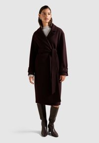 Abrigo envolvente burdeos oscuro con cinturón, con cuello con muescas y mangas largas con puños de botón. Llevado sobre un suéter gris y combinado con botas hasta la rodilla.