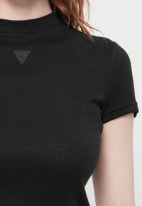 Nærbilde av en person som har på seg en tettsittende svart kortermet topp med et lite prikkete trekantdetalj nær halsen.