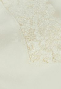 Krämfärgad tyg med en texturerad spetspanel; slät satinytan kompletteras av intrikata spetsdetaljer.