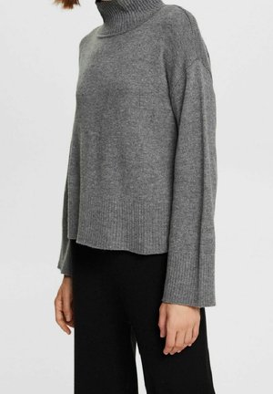 Femme portant un pull à col roulé côtelé gris à manches larges et un pantalon noir sur un fond clair uni.