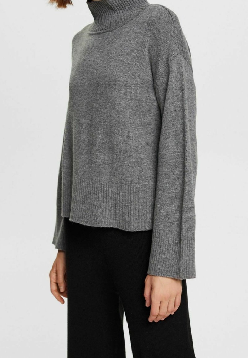 Femme portant un pull à col roulé côtelé gris à manches larges et un pantalon noir sur un fond clair uni.