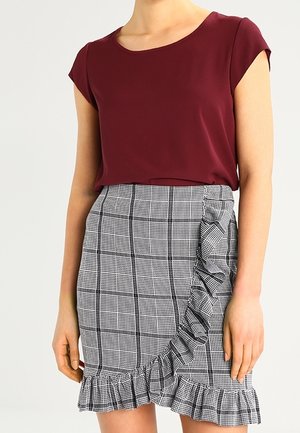 Kobieta w bordowej bluzce z krótkim rękawem i szarej kratkowanej spódnicy z falbanami wzdłuż boku i dołu.