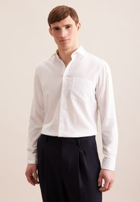 Camicia bianca a maniche lunghe con colletto a punta, tasca sul petto, e una texture liscia, abbinata a pantaloni scuri su misura.
