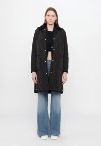 Zwarte gewatteerde jas met capuchon, drukknopen aan de voorkant en zijzakken, gecombineerd met uitlopende denim jeans en witte schoenen.