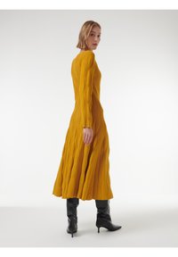 Langermige, mosterdgele gebreide jurk met ribbeltextuur en golvend ontwerp. Gecombineerd met zwarte hoge hakken, vanuit een lichte hoek gepresenteerd.