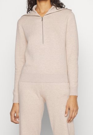 Frau trägt einen beigen Kapuzenpullover mit Reißverschluss und passende Hose, steht mit entspannten Armen seitlich am Körper vor einem einfarbigen Hintergrund.