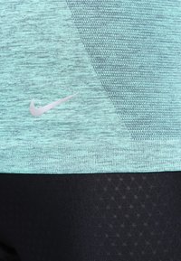 Turkusowa koszulka sportowa Nike ma teksturowany, melanżowy wzór; zestawiona z czarnymi legginsami z geometrycznym wzorem i gładkim materiałem.