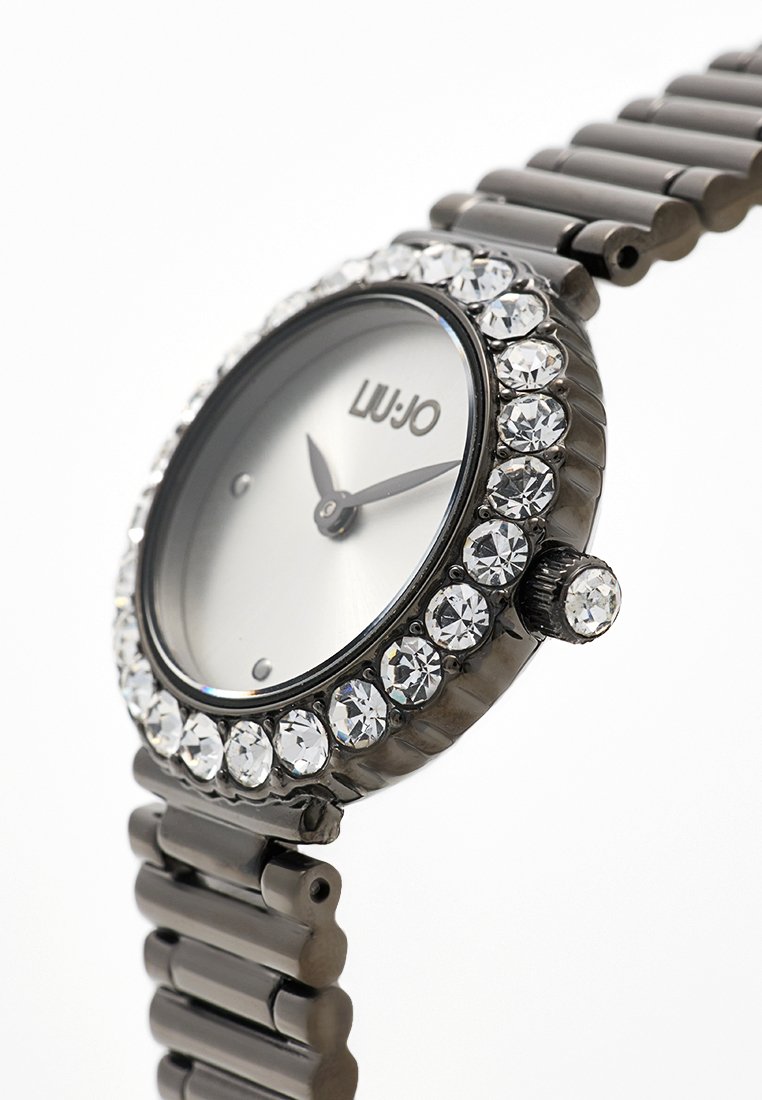 Runde silberne Uhr mit einem mit Kristallen besetzten Gehäuse, grauem Metallarmband und schlichtem Zifferblatt mit schwarzen Zeigern und Logo.