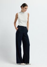 Vrouw in een witte mouwloze top en hoge zwarte wijde broek met haar handen in de zakken, kijkend naar beneden, tegen een effen achtergrond.