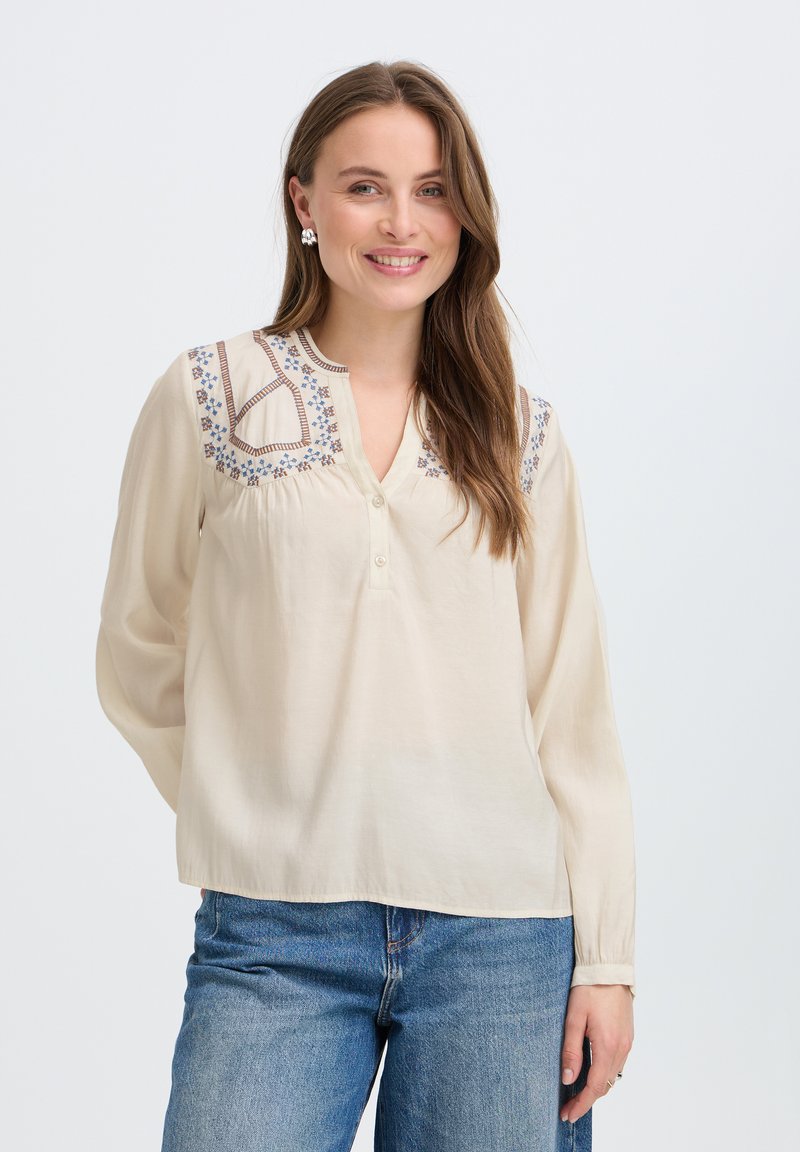 Femme souriante, portant un chemisier crème avec broderies bleues aux épaules, manches longues, jean bleu et boucles d’oreilles argentées, debout sur fond blanc.