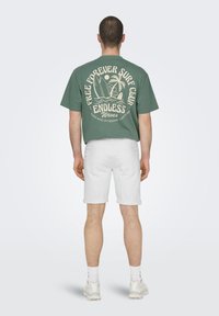 Camiseta de algodón verde con un gran diseño gráfico blanco en la parte posterior que presenta tablas de surf, palmeras y el texto "CLUB DE SURF LIBRE PARA SIEMPRE OLAS INFINITAS". Llevada con pantalones cortos blancos y zapatillas.