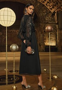 Čierne šaty s priesvitnými rukávmi, ktoré sú ozdobené striebornými detailmi, spárované s metalickými sandálmi na vysokom opätku a metalickou kabelkou. Pozadie s dekoráciou.