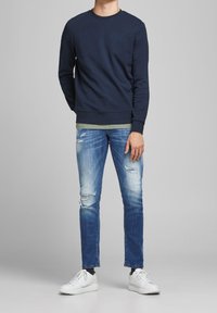 Marinblå crewneck-tröja i kombination med en olivgrön t-shirt, slitna blå jeans och vita sneakers. Avslappnad passform, mjukt tyg, minimal design.