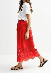 Jupe longue en coton rouge avec des détails découpés floraux à l'ourlet, associée à un t-shirt blanc et des sandales plates noires. Tenue décontractée, vue de côté.