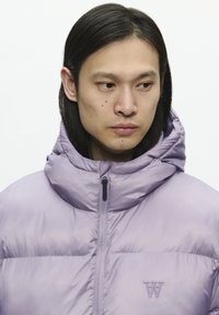 Chaqueta plumífero morada con cuello alto, cierre frontal y logotipo bordado en el pecho. Textura suave y brillante con un diseño ligeramente acolchado.