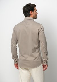 Camisa de manga larga color beige, hecha de un material suave, que presenta un cuello clásico y puños abotonados con botones oscuros distintivos en cada manga.