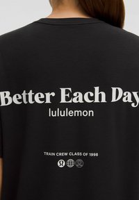 Fekete póló hátulján fehér felirattal: "Jobb minden nap" és "lululemon", valamint "Edző Osztály 1998" és alatta három kis kör alakú logó.