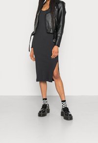 Czarna sukienka z prążkowanej tkaniny z bocznym rozcięciem, zestawiona z krótką czarną skórzaną kurtką i czarnymi platformami. Kraciaste skarpetki stanowią kontrast.