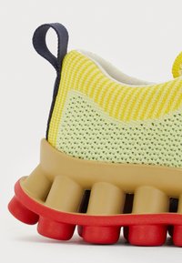 Sapatilha desportiva com um cabedal tricotado amarelo e verde, uma entressola de borracha bege e uma sola exterior segmentada vermelha com detalhes redondos.