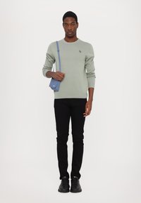 Suéter de malha verde claro, jeans slim-fit pretos e ténis pretos. Uma pequena mala cruzada azul é usada sobre o ombro. Detalhe de logo minimalista no suéter.