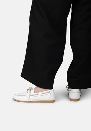 Persona que lleva zapatos estilo mocasín de cuero blanco con cordones beige y suelas marrón claro, combinados con pantalones negros de pierna ancha.