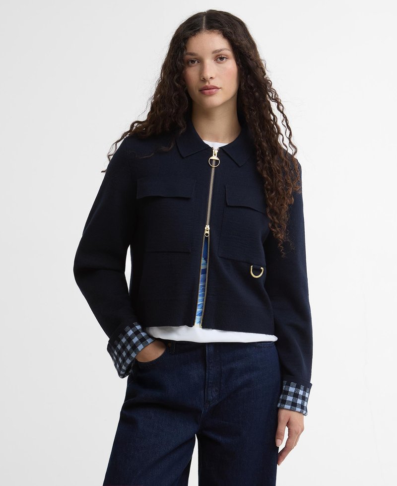 Γυναίκα με μακριά σγουρά μαλλιά φοράει ένα μπλε ναυτικό μπουφάν με φερμουάρ, τσέπες με καπάκι και λεπτομέρειες με καρό μανσέτες, συνδυασμένο με σκούρο τζιν και λευκό πουκάμισο.