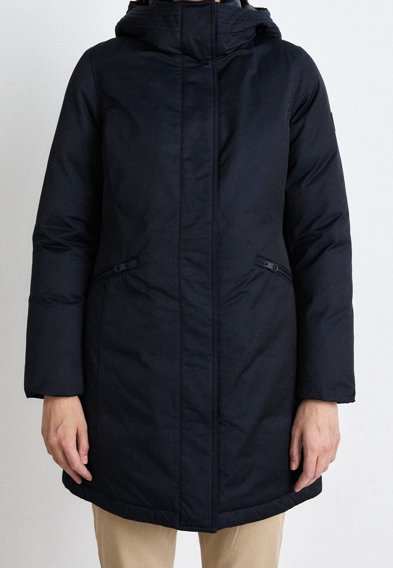 Personne portant un manteau d'hiver noir à capuche avec des poches latérales zippées et un pantalon beige, debout devant un fond blanc uni.