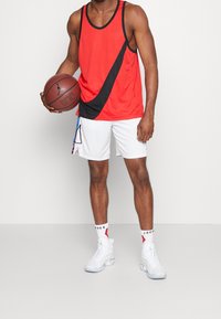 Débardeur rouge avec rayure diagonale noire, shorts de basket blancs ornés de motifs abstraits colorés, et chaussures de sport blanches. Tenant un ballon de basket.