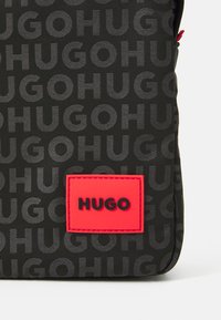 Tissu noir avec un texte "HUGO" en gris foncé répété et un patch rectangulaire rouge en relief portant "HUGO" en lettres noires.