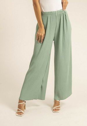 Femme portant un pantalon large côtelé vert clair et des sandales blanches à talons hauts à brides, tenant l'élastique de la taille sur un fond neutre.