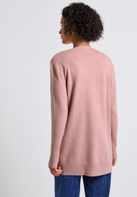 Cardigan tricoté rose, coupe décontractée, poignets et ourlet côtelés, tissu texturé, longueur mi-longue, porté sur un pantalon en denim bleu, fond uni.