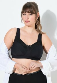 Zwarte beha met kantaccenten op de cups, brede schouderbanden en een strikje in het midden. Het model draagt een losse witte blouse en zwarte broek met polkadots.
