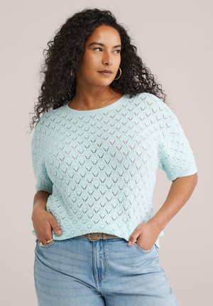 Kvinde med langt krøllet hår iført lyseblå mønstret kortærmet sweater, blå jeans, guldringeøreringe og en tynd halskæde.