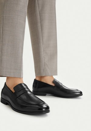 Schwarze Leder-Mokassins, getragen zu beigen karrierten, gekürzten Hosen auf einem schlichten hellen Hintergrund.