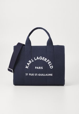 Sac fourre-tout en toile marine avec double poignée supérieure et bandoulière amovible. Comprend un logo blanc imprimé et une adresse, un design structuré et un fond plat.