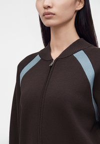 Femme portant un pull zippé brun foncé avec des rayures bleu clair sur les épaules et de longs cheveux noirs.