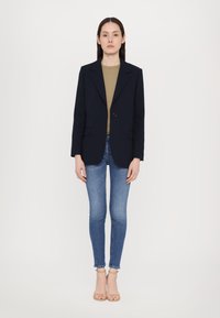 Blazer de marina con un solo botón, solapas con muesca y bolsillos frontales, combinado con una blusa ajustada de color oliva y jeans ajustados azules.