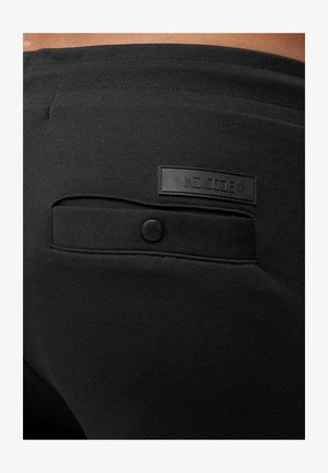 Černé legíny s hladkou texturou a šitým kapsou s knoflíkem. Na pase je viditelné ražené logo "INDICODE".